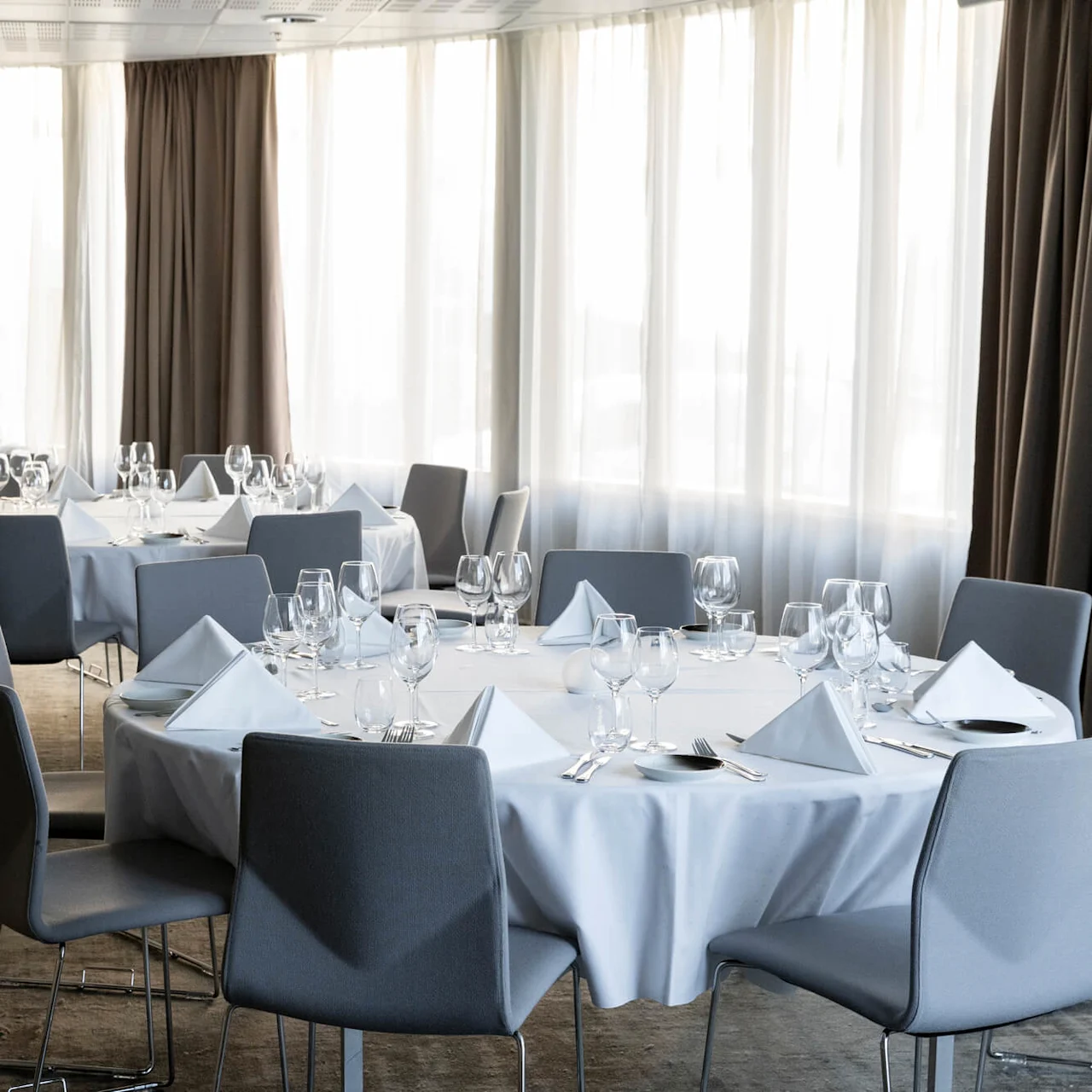Round tables are set elegantly with white tablecloths, napkins, and glassware, surrounded by gray chairs in a softly lit banquet room with large windows draped with curtains.