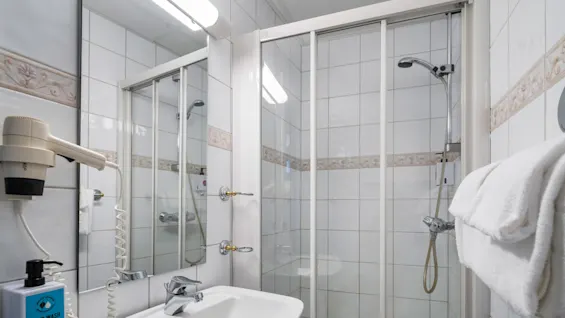 Bathroom with white tiles includes a sink, hairdryer, and mounted soap dispenser labeled "HAND WASH." Adjacent is a shower with a glass door and folded white towels nearby.