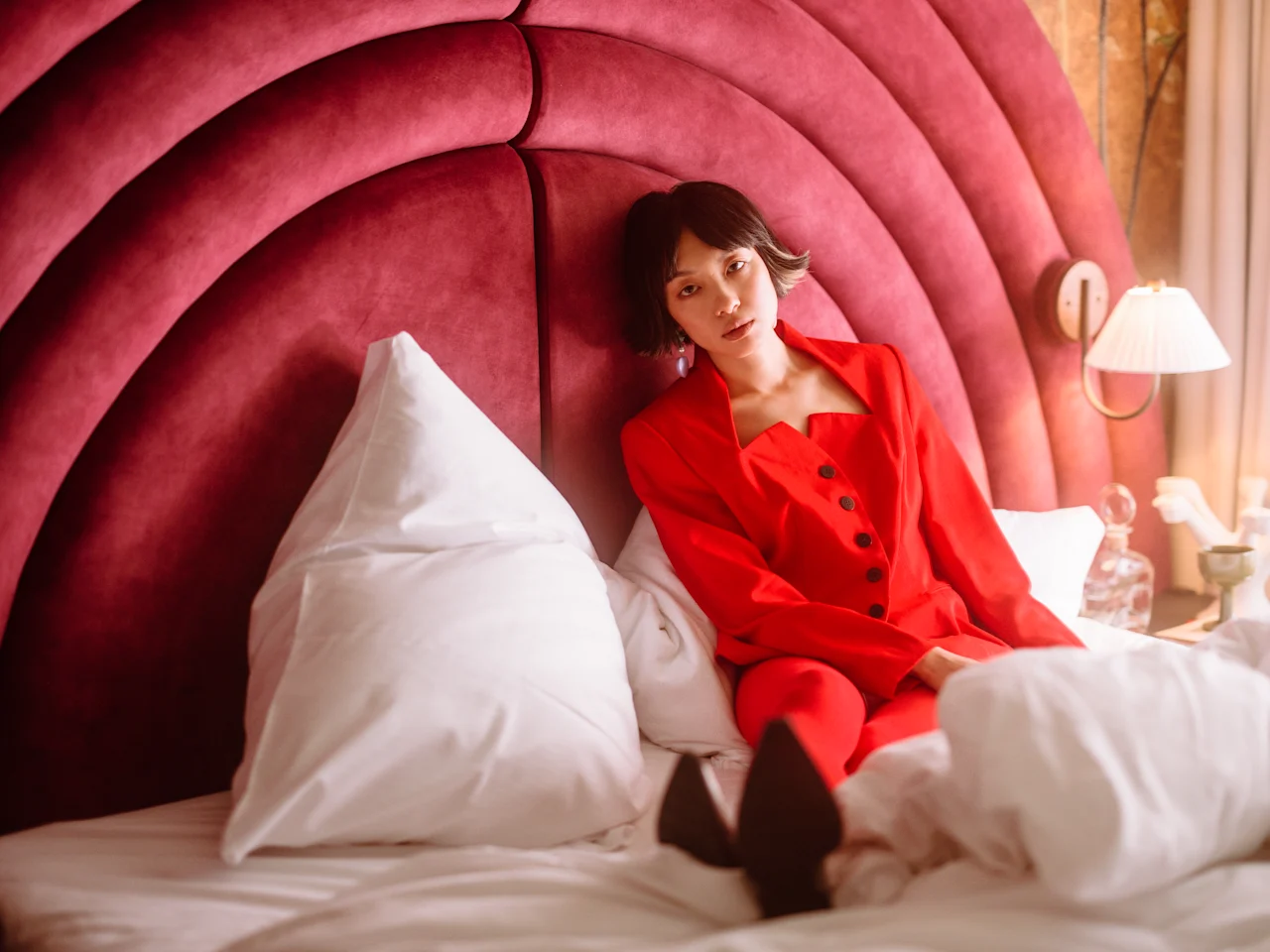 A person in a red outfit sits on a bed against a plush, curved headboard. White pillows surround them. A bedside lamp glows warmly beside the bed.