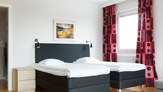 Twin beds are neatly made with white linens in a bright room. A painting hangs above the headboard, and red patterned curtains flank a window, highlighting the wooden floor.