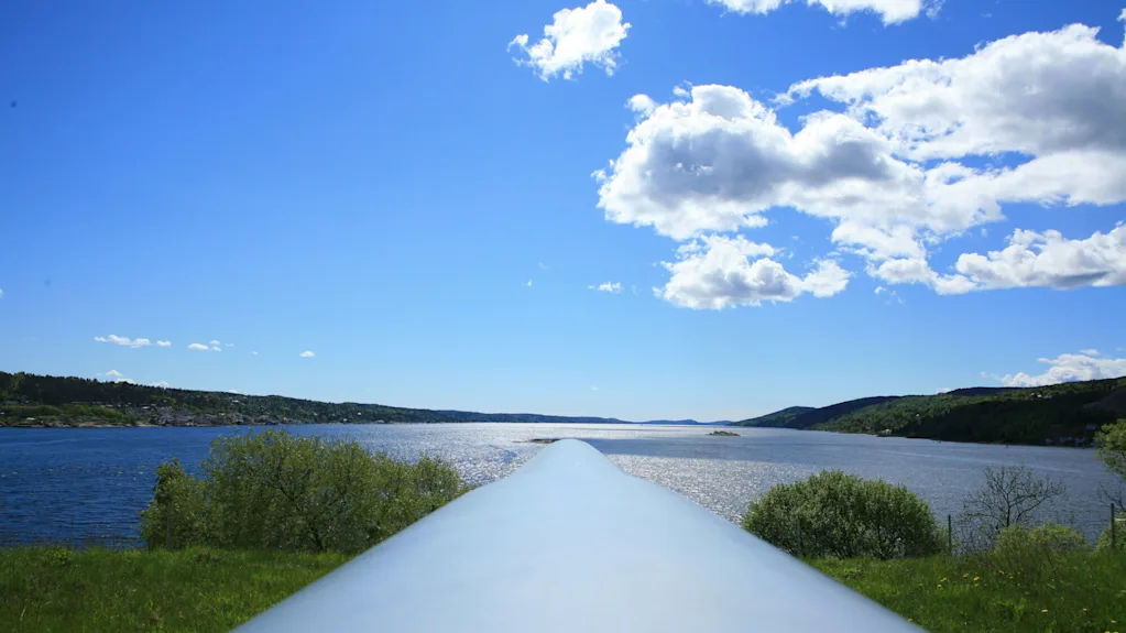 A large metallic pipe extends into a scenic landscape featuring a tranquil river surrounded by lush green hills and scattered clouds overhead under a bright blue sky.