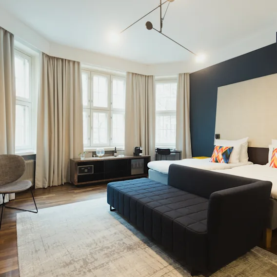 Moderni hotellihuone, jossa on kaksi sänkyä, tumma sohva, nojatuoli ja suuret ikkunat vaaleilla verhoilla.