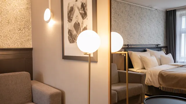 Et koselig soverom med en beige sofa, opplyste stålamper og innrammet kunstverk. En pent oppredd seng med hvitt sengetøy ved siden av et vindu med lyse og mørke gardiner.