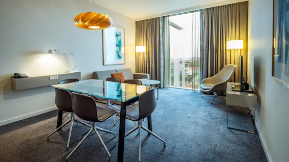 Ett modernt vardagsrum har ett glasbord med stolar, en soffa och en fåtölj. Stora fönster med gardiner erbjuder stadsutsikt. Hyllor och lampor kompletterar inredningen.
