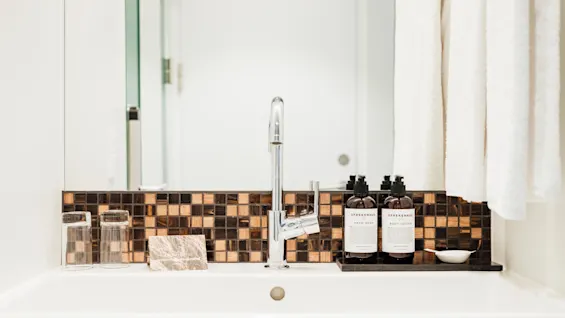 En hvid badeværelsesvask med en skinnende kromhane under et spejl, har to glas og brune mosaikfliser, samt flasker mærket "SPREKENHUS HAND WASH" og "BODY LOTION."