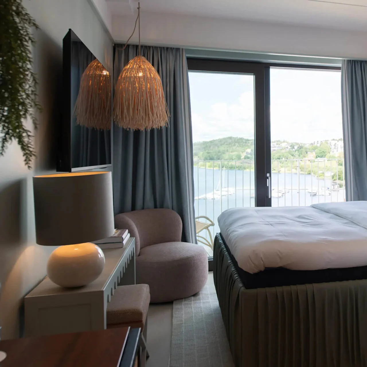 A neatly made bed faces a large window with a scenic lake view. Cozy seating, modern lamps, and a television complement the serene, stylish room decor.