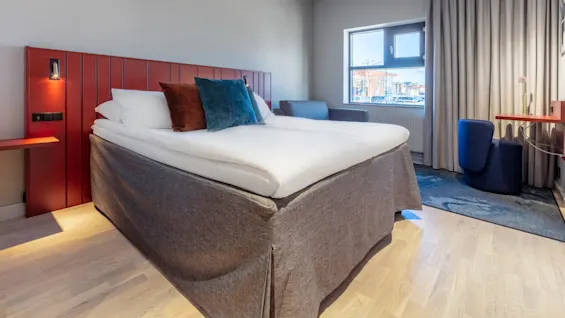 A neatly made bed with pillows against a red headboard, in a bright, modern bedroom with a window and curtains.
