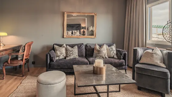A plush sofa with patterned cushions sits in a living room. A coffee table with a candle is centered. Wooden desk and chair are on the left, window on the right.