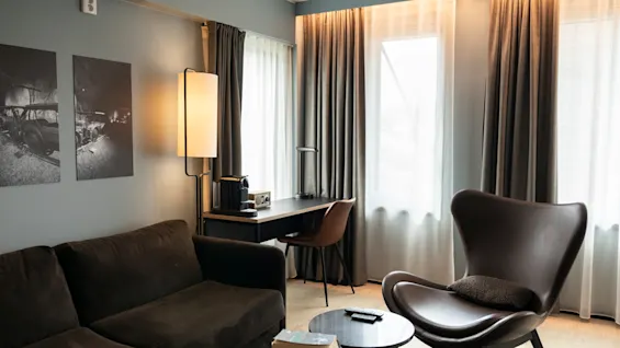 Et koselig hotellrom med mørkebrun sofa, skinnlenestol, skrivebord med kaffemaskin og svart-hvitt kunstverk på veggen.
