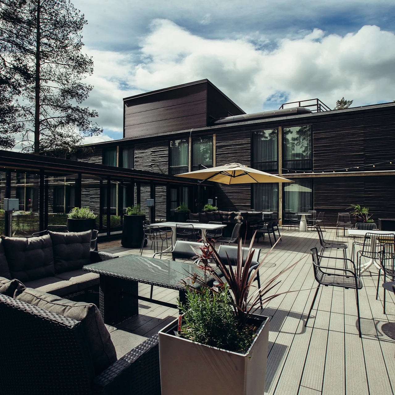 Modern outdoor patio with wicker sofas and glass tables arranged under a large yellow umbrella. Surrounded by tall trees, the dark wood building stands in the background against a cloudy sky.