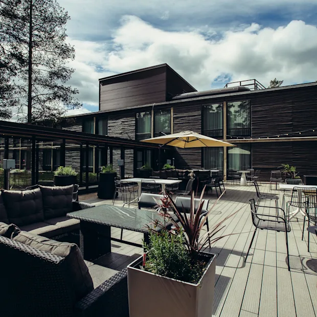 Moderni ulkoterassi, jossa on rottinkisohvia ja lasipöytiä suuren keltaisen aurinkovarjon alla. Tumma puinen rakennus ja korkeat puut ympäröivät aluetta pilvisen taivaan alla.