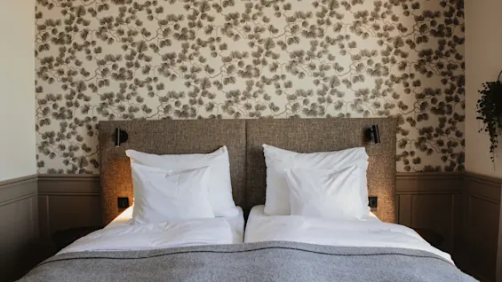 Ett mysigt hotellrum med en säng bäddad med vita lakan och en grå filt, mot en mönstrad tapet.
