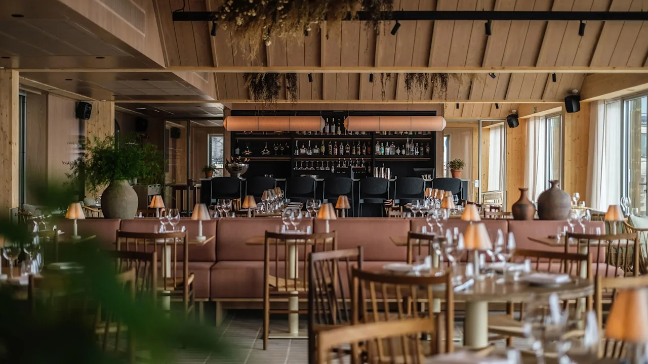 Moderne restaurant fyldt med træborde og stole, pyntet med vinglas og små lamper, vender mod en fuldt udstyret bar. Store vinduer og potteplanter skaber en hyggelig, indbydende atmosfære.
