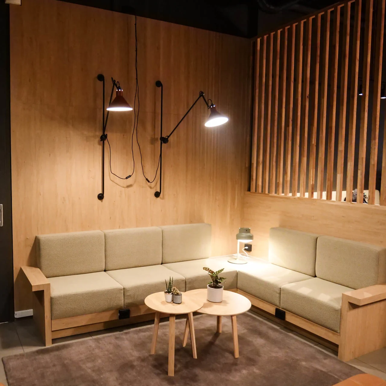 A cozy seating area features two beige sofas forming an L-shape, a small round wooden table with potted plants, and wall lamps, all set against light wood paneling. 