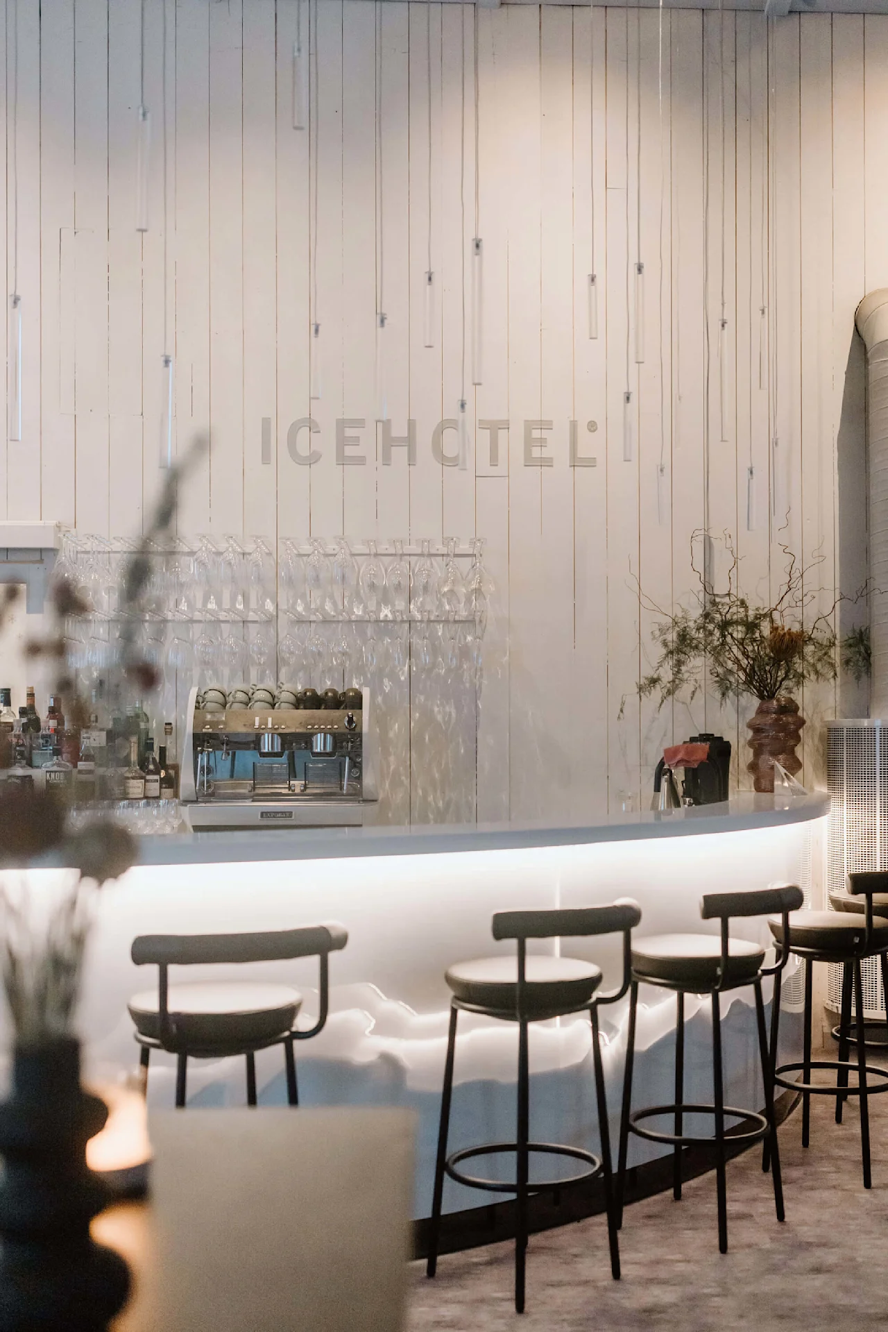 En buet bardisk viser stole og glas i et minimalistisk rum med blød belysning. "ICEHOTEL"-tekst er monteret på væggen blandt hængende dekorationer.