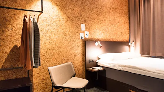 A cozy room features a chair beside a bed with a cork-textured wall. Clothes hang on a rack, and light shines on a small shelf with a notebook.