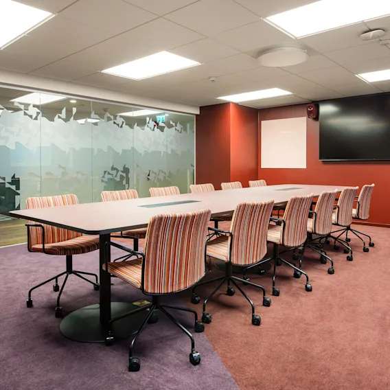 A long conference table with striped chairs is set in a modern meeting room. A large screen and frosted glass wall create a professional environment.