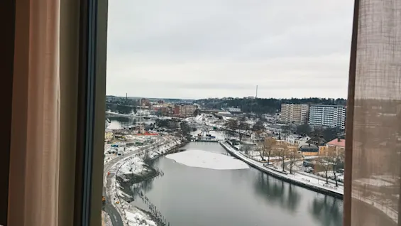 En högvinkelvy från ett fönster visar en vinterstad med en flod, snötäckta stränder och byggnader under en molnig himmel.