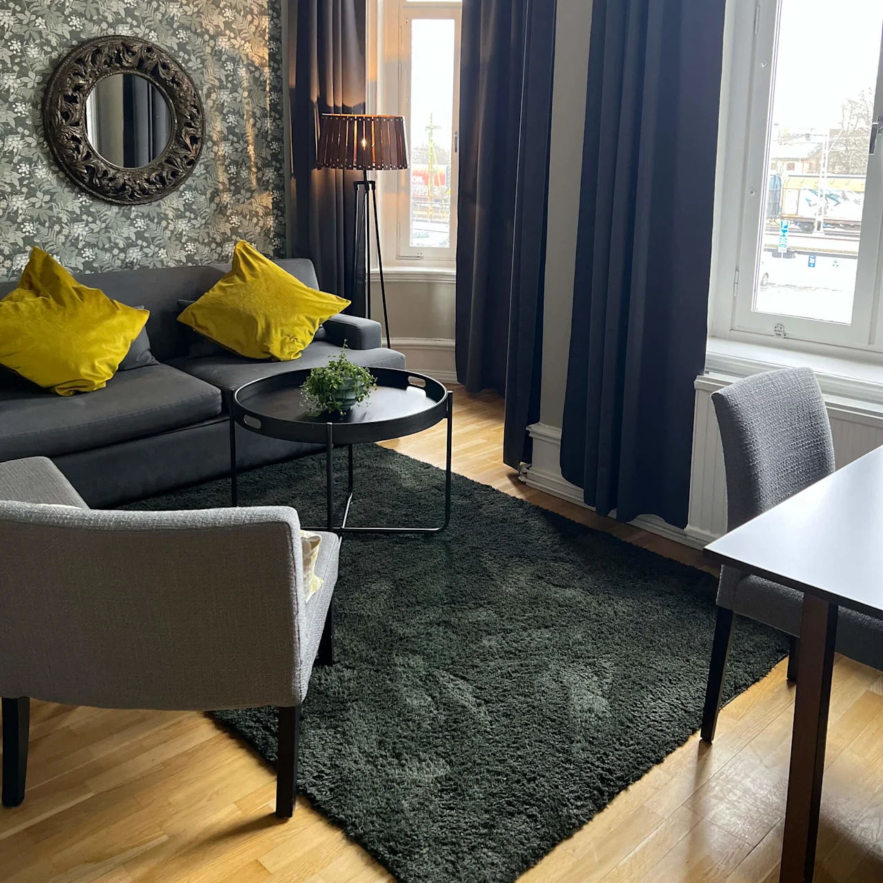 A cozy living room with modern decor, featuring a gray sofa with yellow pillows, and large windows draped in dark curtains.