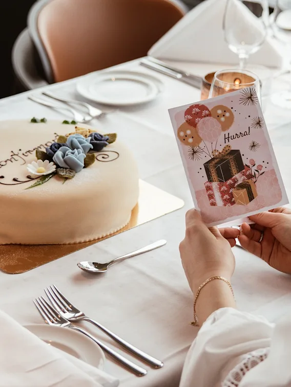 En person leser et gratulasjonskort med 'Hurra' ved et dekket bord med en dekorert kake, som indikerer en feiring. 🎈