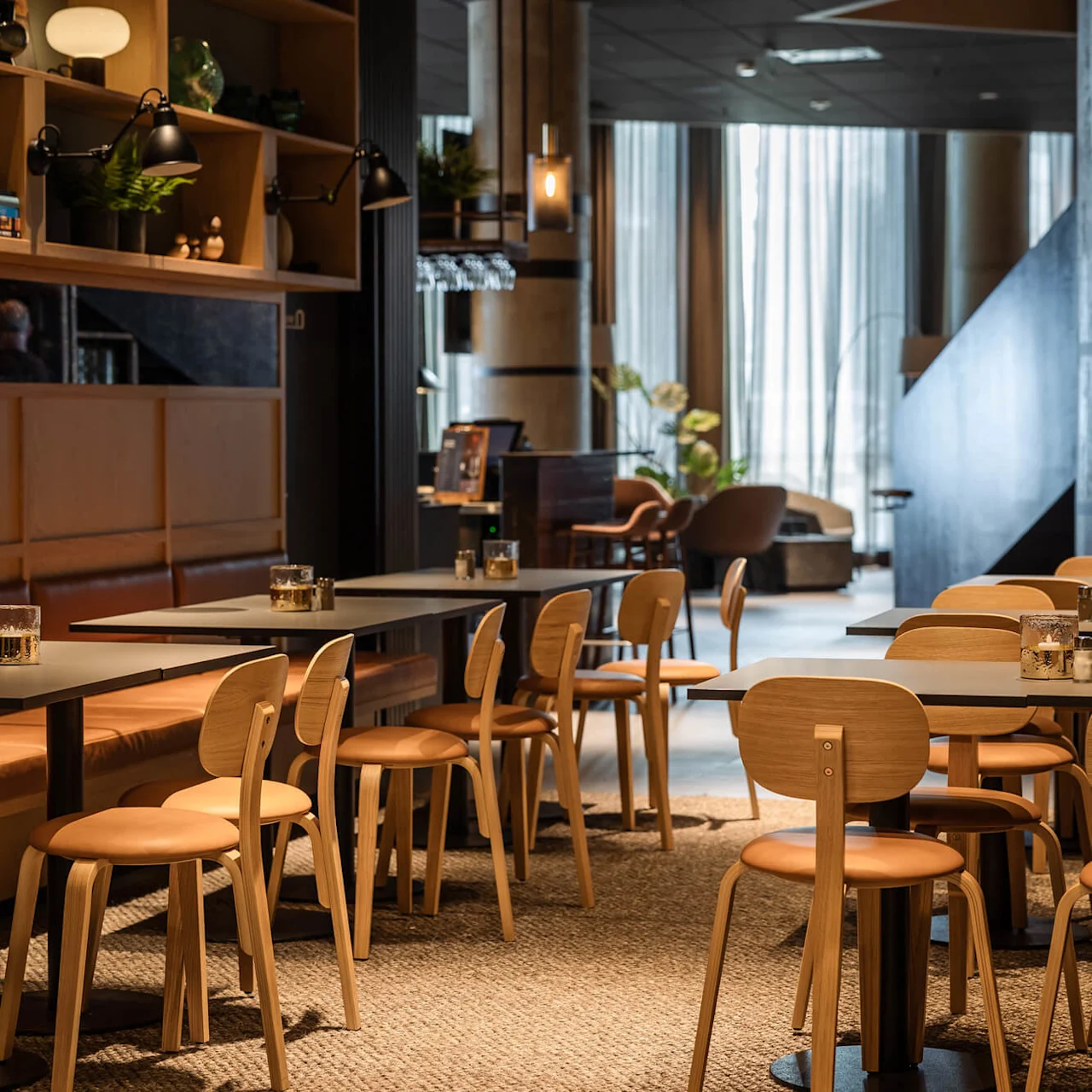 Borde med stole, pænt arrangeret i en hyggelig café med varm belysning. Træreoler viser dekorative genstande, mens store vinduer lader naturligt lys strømme ind. Gulvtæppe understøtter den indbydende atmosfære.