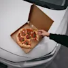 En hånd rækker ud efter et stykke pepperoni-pizza fra en æske, der hviler på en bilhjelm, et praktisk stopover-måltid på Hotel Falkenberg.