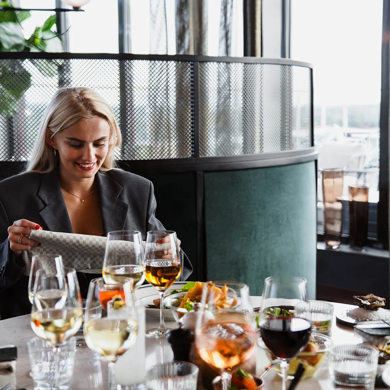 En kvinde smiler, mens hun åbner en gave ved et spisebord, omgivet af vinglas og tallerkener med mad i en hyggelig restaurantindstilling.