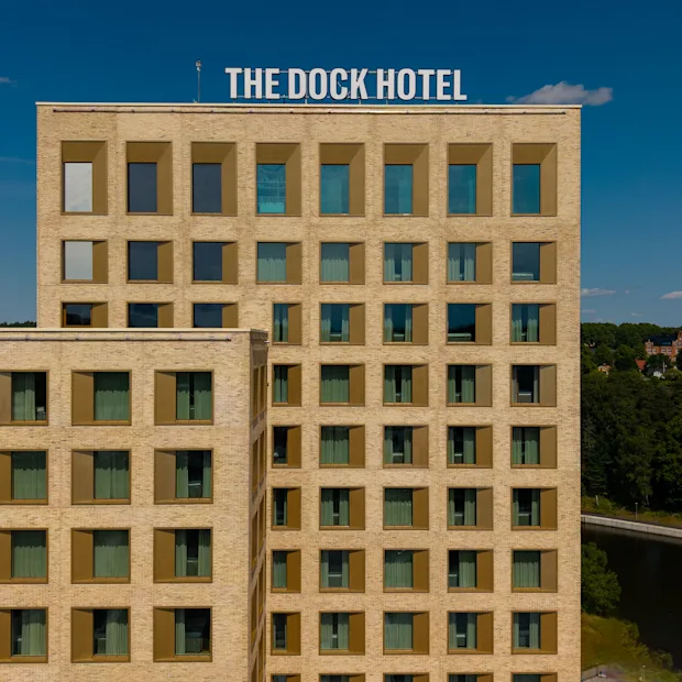 The Dock Hotel, en moderne bygning med mange vinduer, står mot en blå himmel med en grønn skogkledd bakgrunn.
