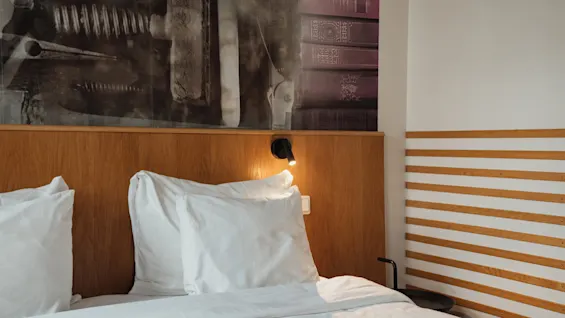 A cozy hotel room bed at Home Hotel Uppsala with white linens, a wooden headboard, and a unique mural of books and machinery.
