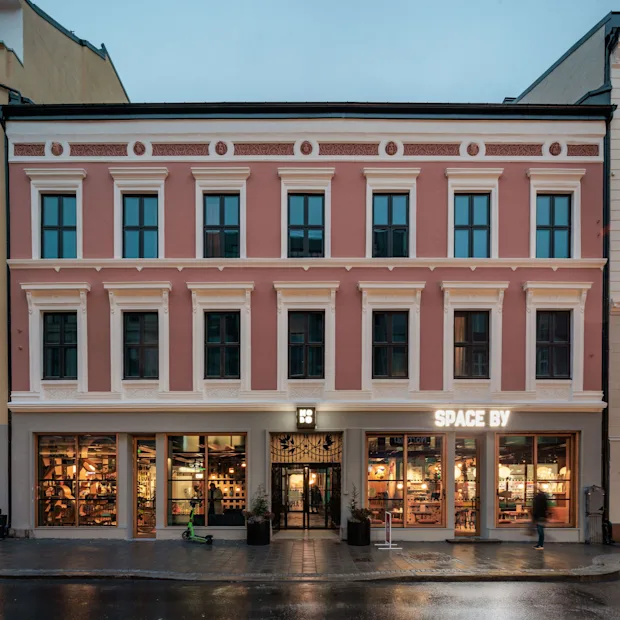 A pink building with white trim and many windows. The ground floor has large illuminated display windows and an entrance. Text reads SPACE BY.