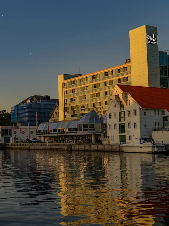 A large hotel building with "QUALITY" sign overlooks a waterside area at sunset. Boats are docked near the shoreline, and a curved bridge arches across the water.