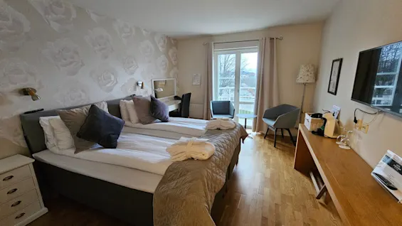 A bright and cozy hotel room with a double bed, wooden floors, and a glass door leading to a balcony