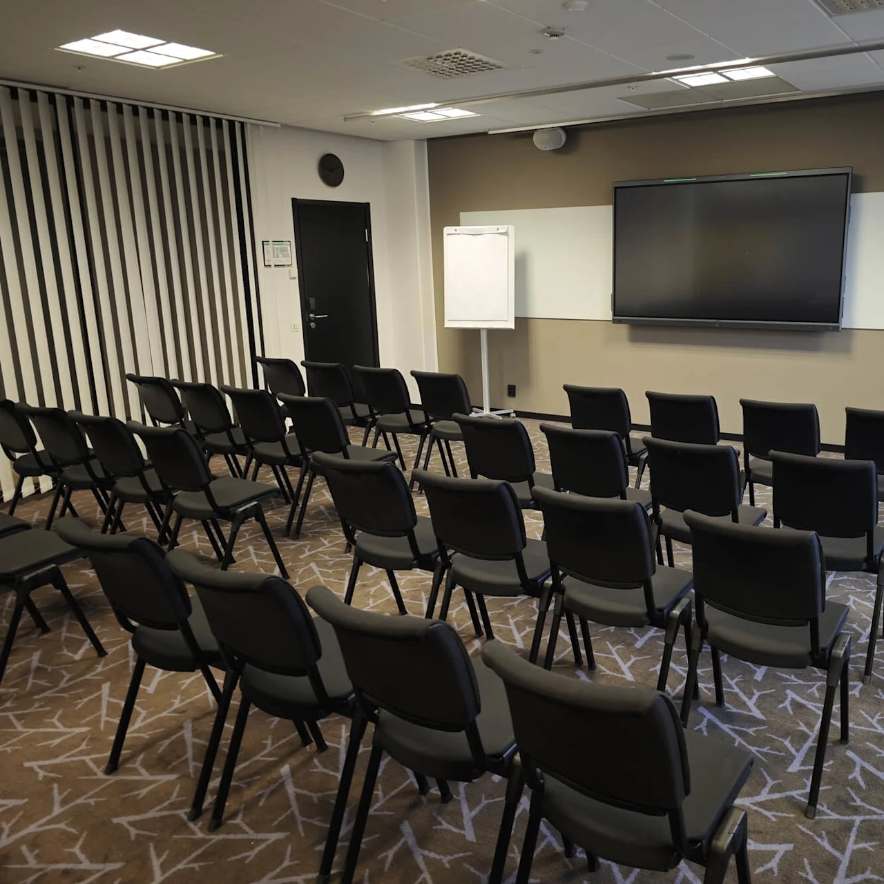 Stolar är placerade i rader vända mot en tom skärm och whiteboard i ett konferensrum med matta. Ett blädderblock och en talarstol finns också.