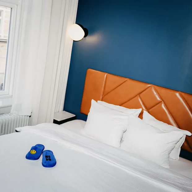 Moderni hotellihuone, jossa on sininen seinä, nahkainen sängynpääty ja siniset tossut sängyllä.