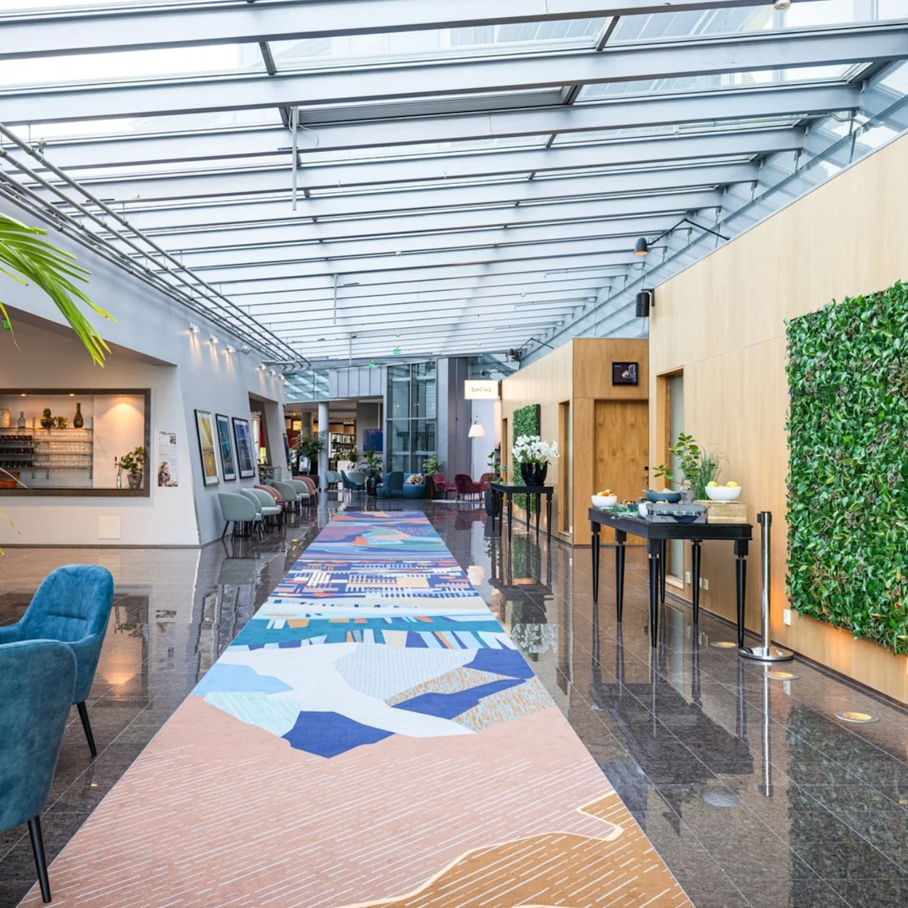 A vibrant hallway features a colorful carpet; lush green plant wall; cozy seating; and a distant lounge area under a glass ceiling, creating an open, sophisticated atmosphere.