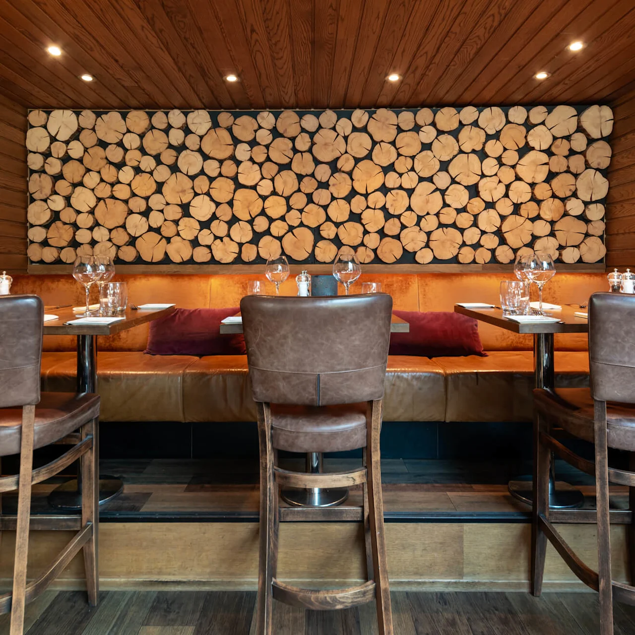 Matbord är uppställda med glas och tallrikar, arrangerade runt läderstolar och bänkar. En träväggsdesign med stockar dekorerar bakgrunden i en varm upplyst restaurang.