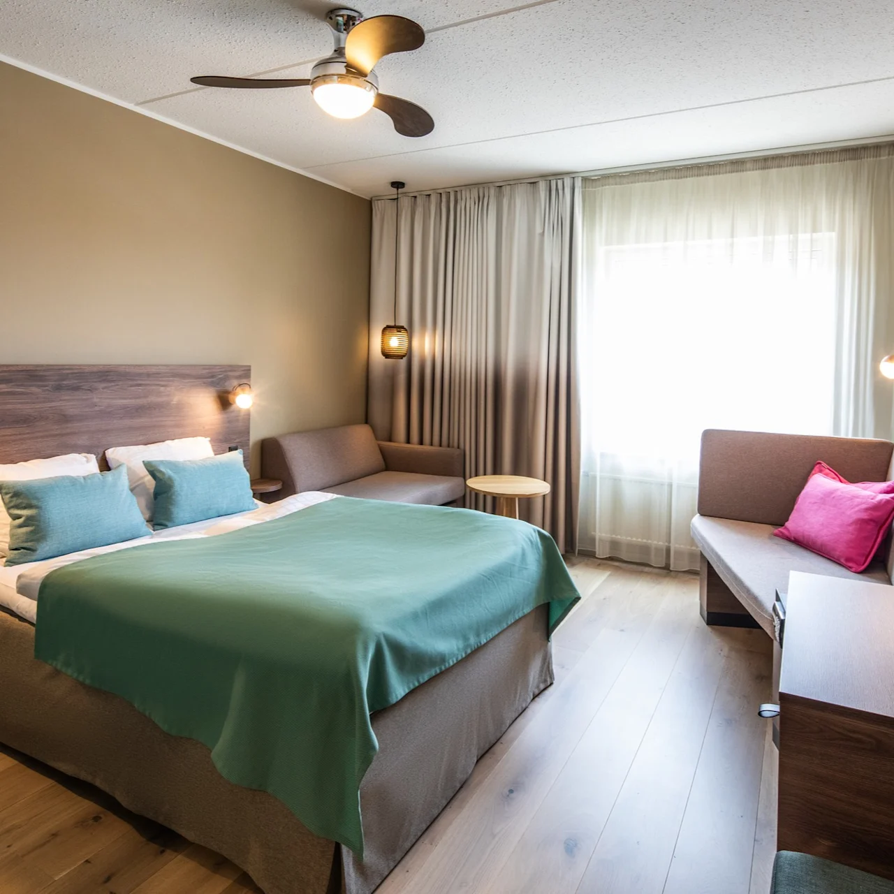 Bed positioned centrally with teal bedding; wall-mounted lamps illuminate both sides. Nearby, a couch accessorized with a pink cushion sits under sheer-draped window. Wooden floor and ceiling fan complete the cozy room.