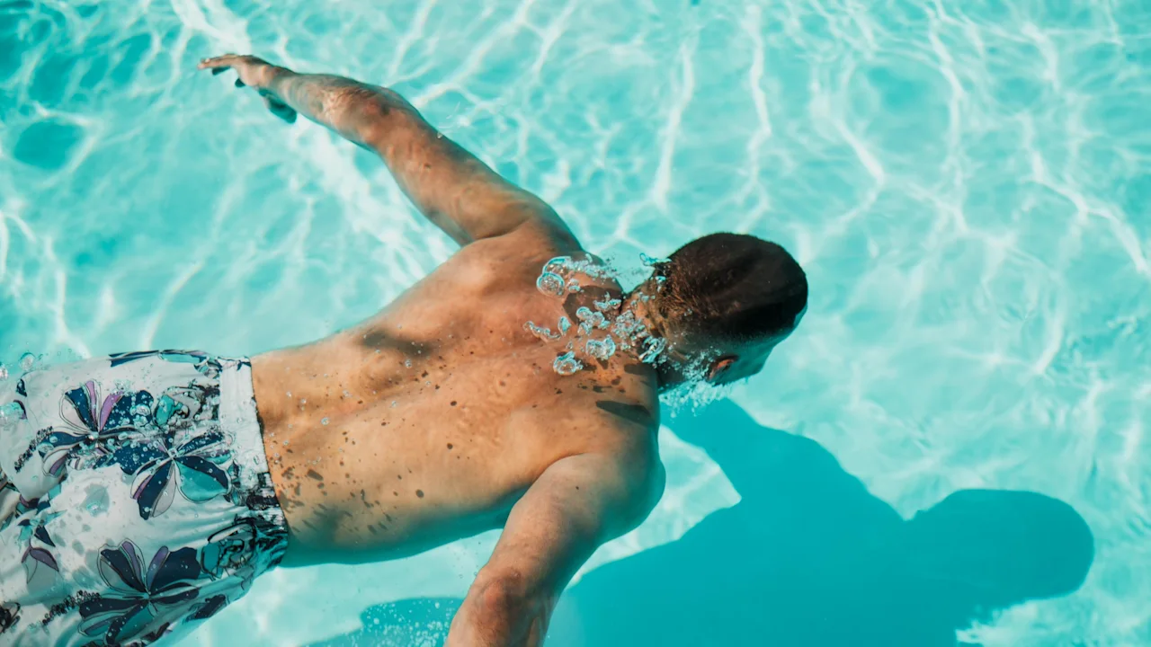 En person simmar under vattnet i en klarblå pool, med solljus som glittrar på vattenytan.