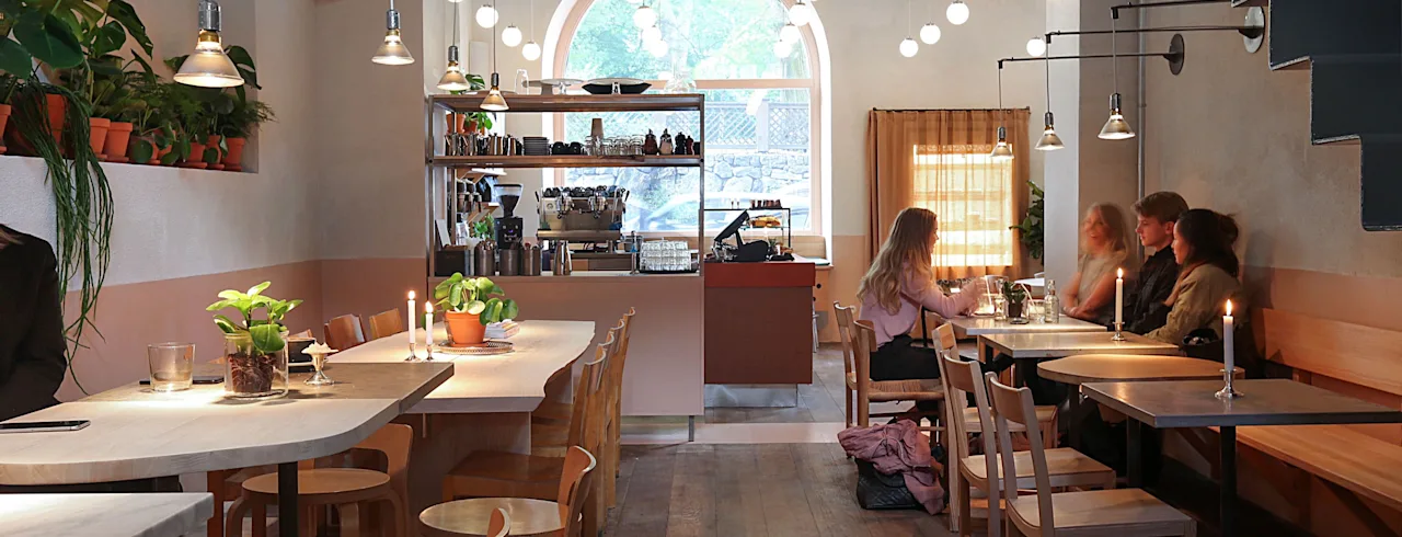 Folk nyder et måltid eller drikke på en hyggelig café med varm belysning, planter og et stort buet vindue.