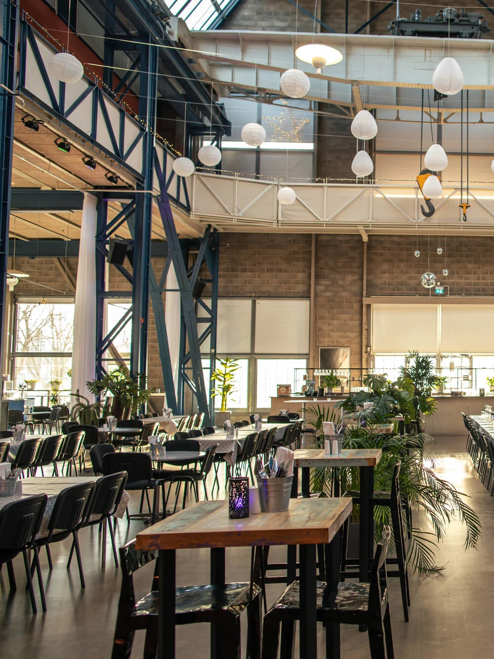 Bord är uppställda med stolar och bestick, i rader under hängande lampor i en stor, industriell hall med högt tak och växter.