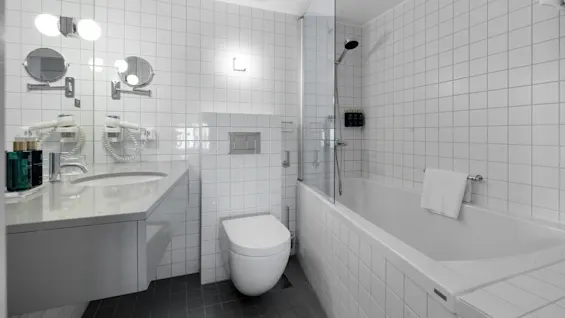 Et moderne bad har et veggmontert toalett og servant, samt et badekar med en glasskjerm. Hvite fliser dekker veggene og reflekterer sterkt lys, og skaper en ren atmosfære.