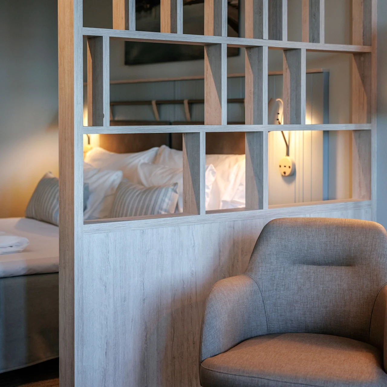 A beige armchair sits beside a wooden partition with open sections. Behind it, a neatly made bed with striped cushions is illuminated by a wall lamp, creating a cozy atmosphere.