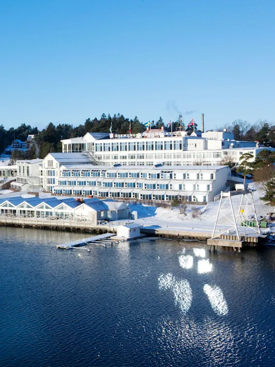 A large white building with the text "Stenungsbaden Yacht Club" stands near a snow-covered shoreline by the water, surrounded by trees under a clear blue sky.