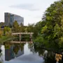 En moderne glasbygning står ved en rolig flod, spændt af en gangbro, alt indrammet af frodigt grønt.