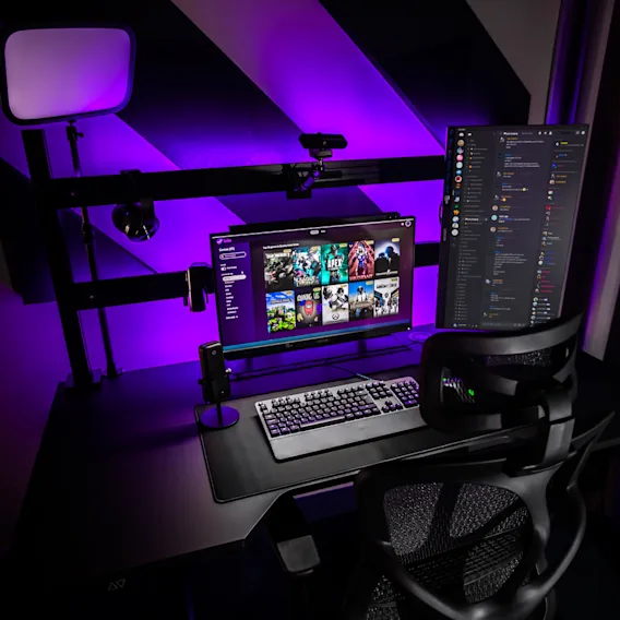 Ett modernt svart skrivbord med dubbla skärmar; en visar ett spelbibliotek med titlar som Apex Legends, Among Us, Minecraft. En lila LED-lampa lyser upp den ergonomiska stolen och omgivningen.
