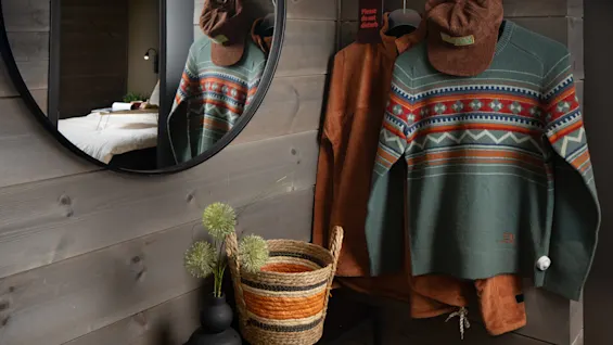 Et rundt spejl reflekterer et hyggeligt rum med en seng; en farverig sweater og hat hænger på kroge under teksten: "Please clean". Et konsolbord har vaser og en flettet kurv.