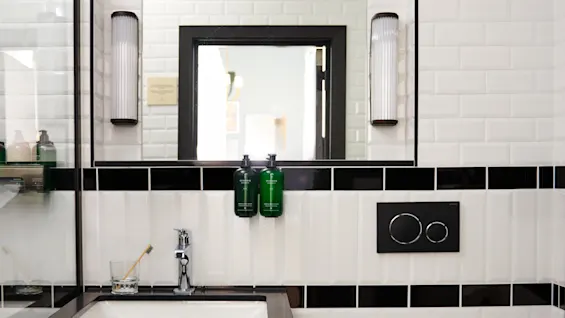 A black sink with faucet sits below a large mirror, flanked by lights, in a white tiled bathroom with black accents. A wall-mounted toilet is adjacent.