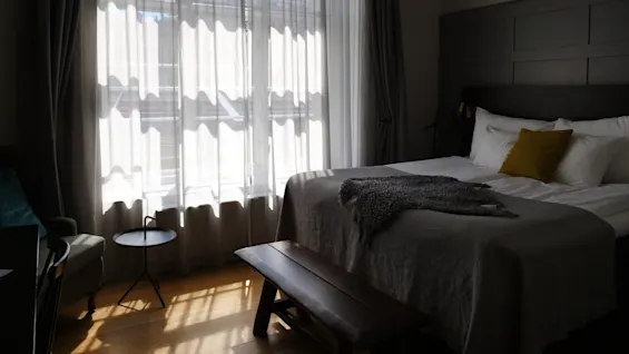 En pænt redt seng med en gul pude og gråt tæppe sidder under sollys, der filtreres gennem gennemsigtige gardiner, med et lille bord og lænestol i nærheden.