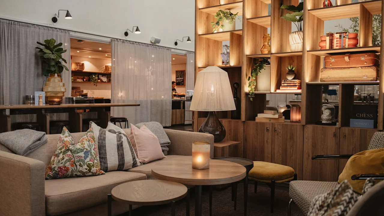 En koselig salong har beige sofaer med fargerike puter, et rundt trebord med et tent lys og en høy hylle med bøker og planter i et svakt opplyst rom.