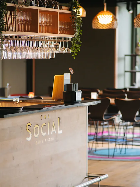 Bardisk dekorerad med glas och gröna växter, placerad i en stilfull bistro med flätade lampor och moderna sittplatser. Text på disken: "THE SOCIAL BAR & BISTRO"